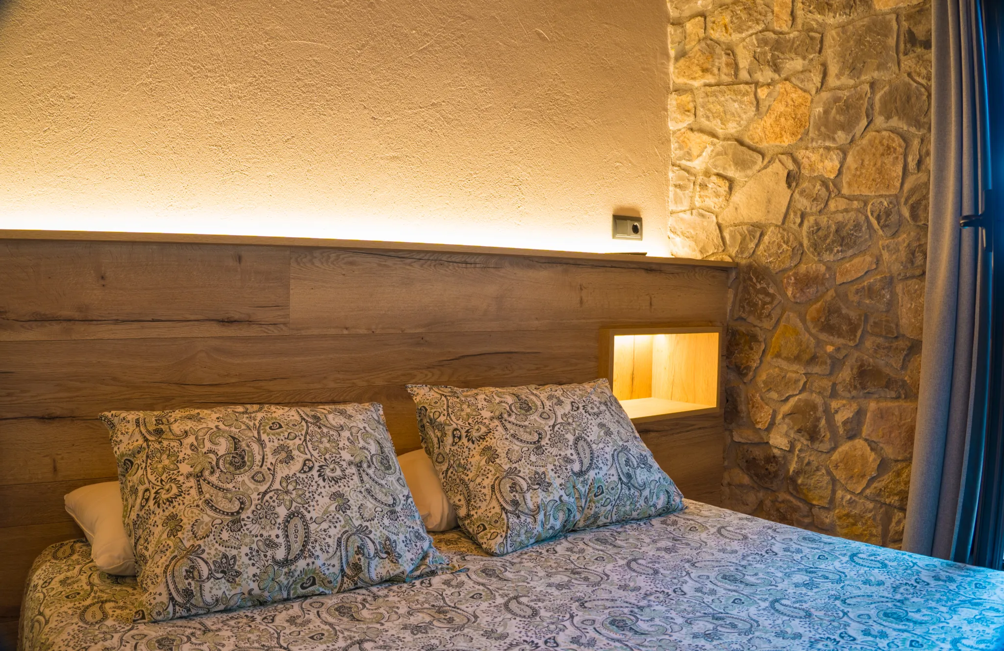 Llit de matrimoni amb capçal de fusta il·luminat i paret de pedra