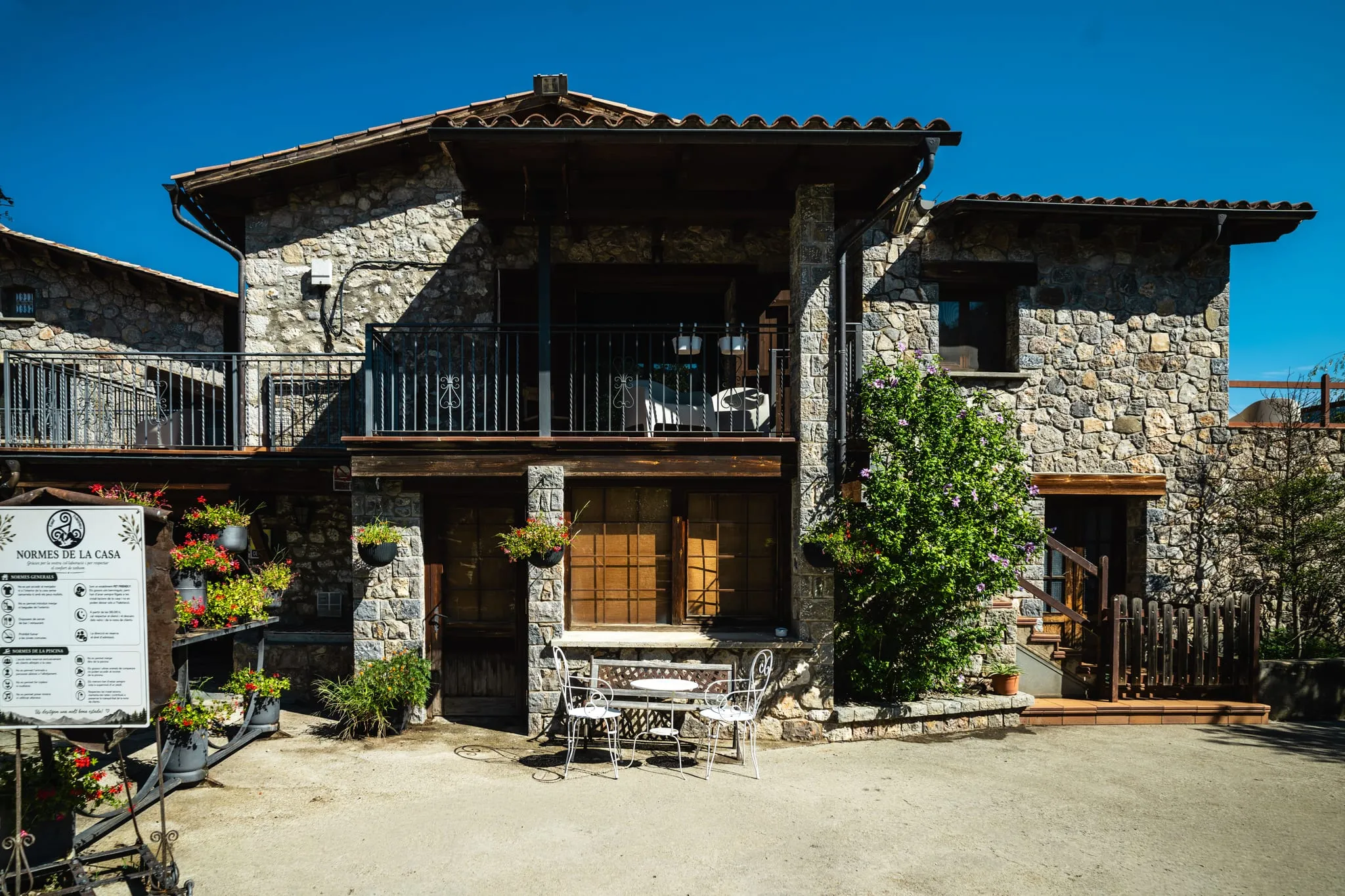 La casa de pedra amb balcó de fusta, testos de flors i una tauleta a l'entrada