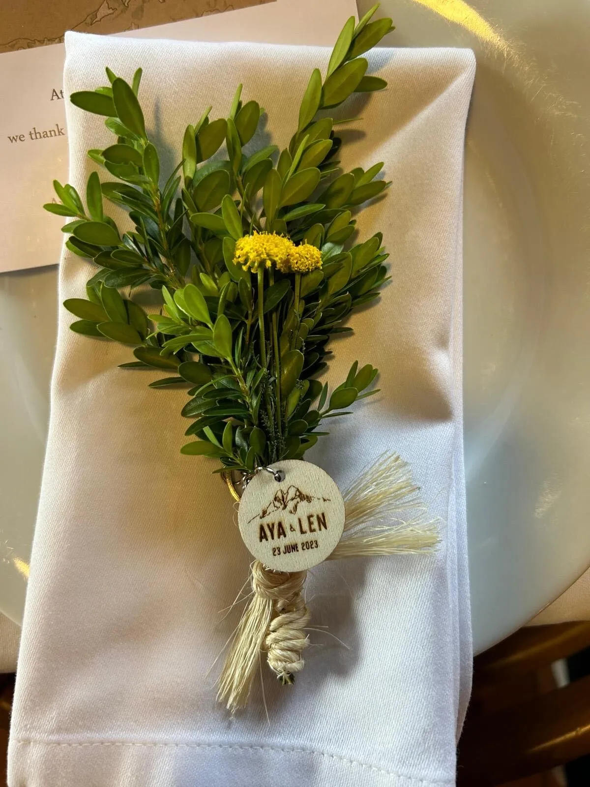 Detall de casament sobre el tovalló: un ram de boix amb flors grogues i una placa de fusta gravada