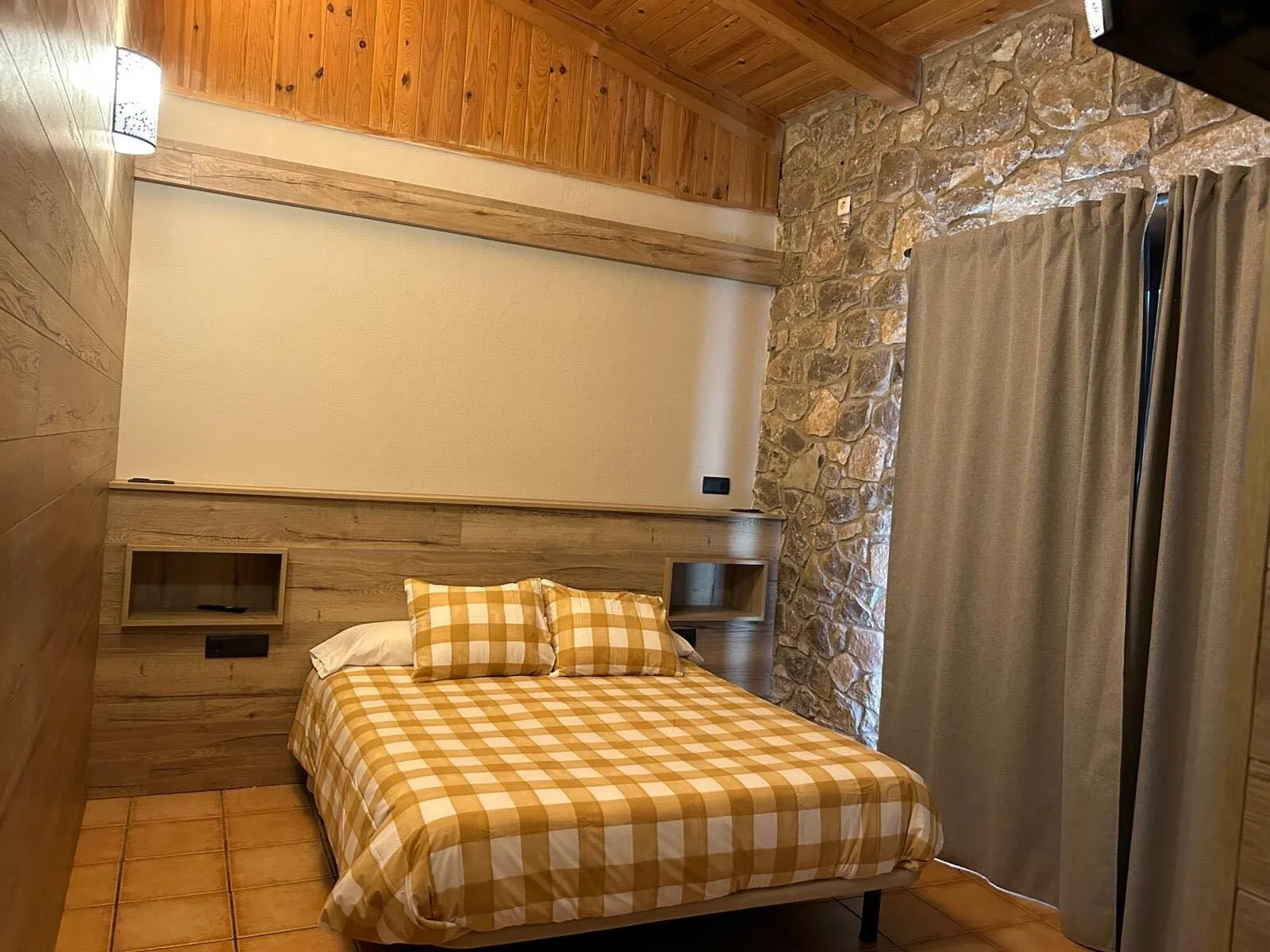 Habitació doble amb paret de pedra, capçal de fusta i cobrellit de quadres