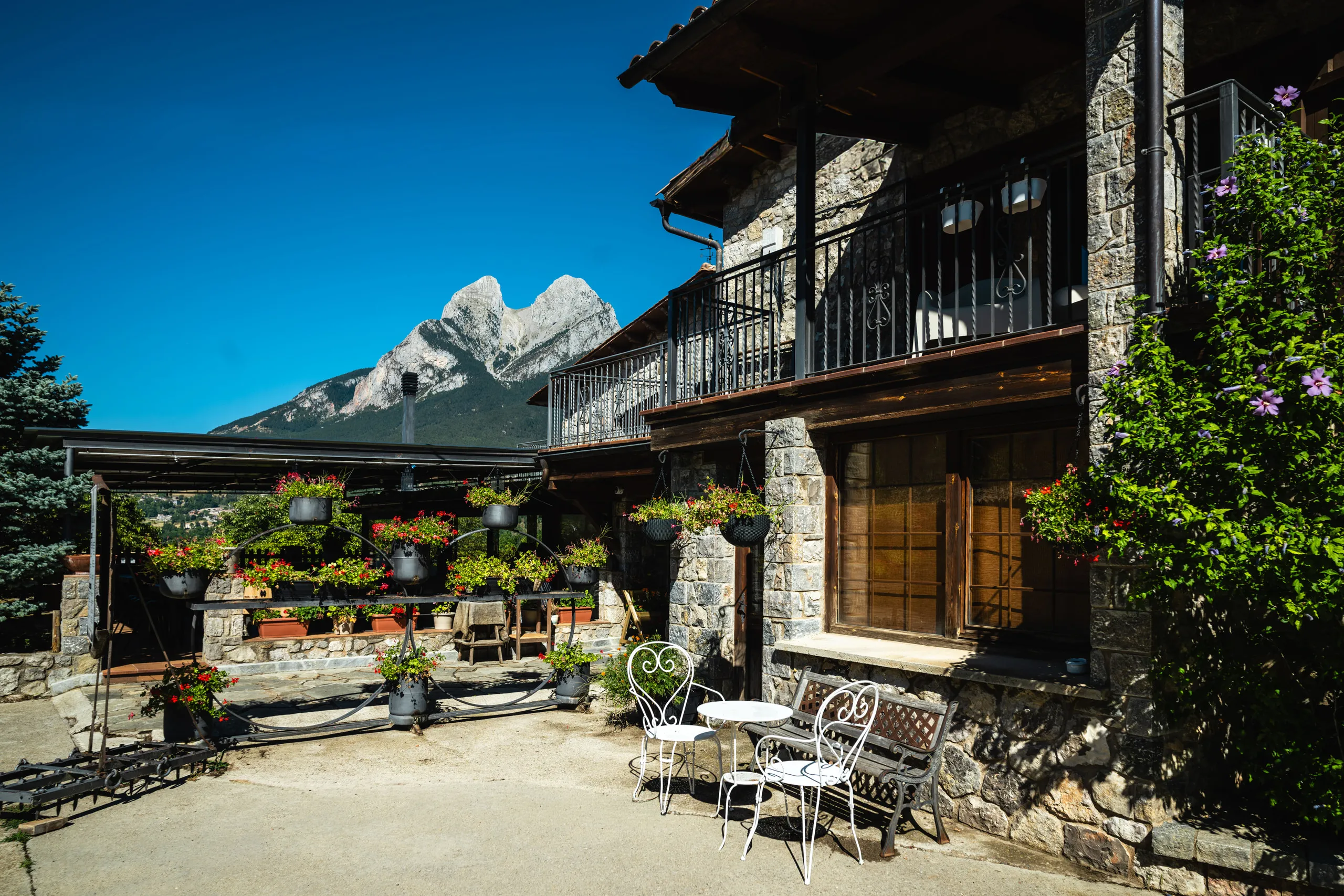 El pati de la casa amb taules de ferro blanques i testos de flors, amb el Pedraforca al fons