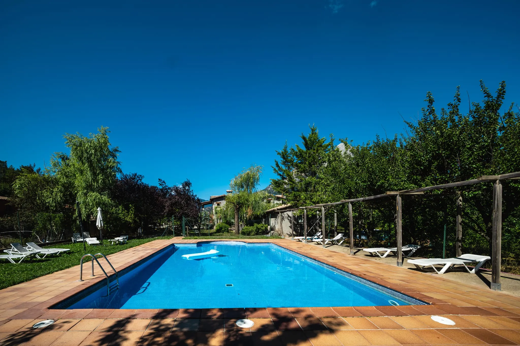 La piscina amb gandules a la gespa, entre arbres i sota un cel blau